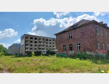 Eine verlassene Gegend mit zwei stark heruntergekommenen Gebäuden: ein mehrstöckiges Betongebäude mit zerstörten Fenstern und Graffiti sowie ein Backsteingebäude mit ähnlichen Schäden. Im Vordergrund wächst unkontrolliert Gras und Vegetation, im Hintergrund ist ein bewölkter Himmel zu sehen.