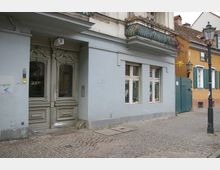 Wohnhausfassade in einer gepflasterten Straße, mit einer dekorativen Doppeltür in grauer Farbe, Fenstern neben der Tür sowie einem Balkon mit schmiedeeisernem Geländer darüber. Rechts daneben schließt ein gelb gestrichenes Gebäude mit grünen Fensterläden an. Lampen im historischen Stil stehen entlang der Straße.