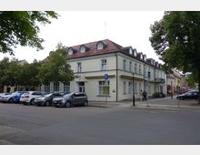Zweistöckiges Gebäude mit hellgelber Fassade und rotem Dach, das mehrere Dachgauben enthält, an einer Straßenecke. Im Vordergrund parkende Autos, Gehweg mit Bäumen und einige Personen; rechts sind Fahrräder und weitere kleine Gebäude sichtbar.