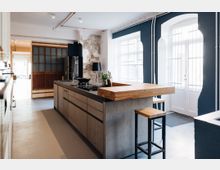 Moderne Küche mit einer zentralen Kücheninsel aus Betonoptik und einer Arbeitsplatte aus Holz. Im Hintergrund sind weiße Wände mit freiliegendem Putz, große Fenster mit Gitter und eine Glas-Holz-Schiebetür sichtbar.