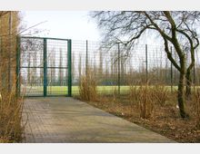 Gepflasterter Weg, der zu einem grünen Gittertor führt, das einen eingezäunten Sportplatz mit Rasen umschließt; umgeben von kahlen Bäumen und Sträuchern. Im Hintergrund sind hochgewachsene Baumreihen sichtbar.