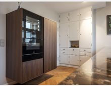 Moderne Küche mit einem Einbauschrank aus dunkelbraunem Holz mit integriertem Backofen und anderen Geräten auf der linken Seite. Rechts stehen weiße Oberschränke mit mehreren Schubladen, Griffen und einer offenen Ablage; der Boden besteht aus Fischgrätenparkett.