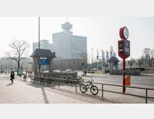 Städtische Straßenszene in Berlin mit der U-Bahn-Station „Theodor-Heuss-Platz“ im Vordergrund, mehreren geparkten Fahrrädern und einem großen Bürogebäude mit moderner Architektur im Hintergrund. Eine Uhr und Werbetafel stehen auf einem roten Mast am Straßenrand.