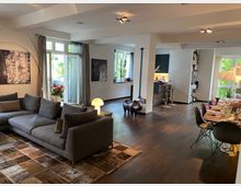 Modernes Wohnzimmer in einer offenen Wohnfläche mit dunklem Holzboden. Sichtbar sind eine graue Couch auf einem gemusterten Teppich, ein Esstisch mit gedeckten Tellern, große Fenster mit Vorhängen, abstrakte Kunstwerke an den Wänden und eine kleine Küchenzeile im Hintergrund.