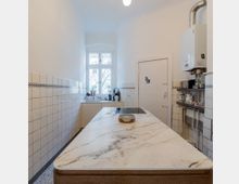 Schmale Küche mit weißen Wandfliesen und schwarzer Bordüre, zentralem Küchenblock mit heller Marmorarbeitsplatte, einem Fenster im Hintergrund und einer Gastherme an der rechten Wand.