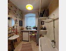 Kleines Badezimmer mit Badewanne, beiger Fliesenwand und Duschvorhang mit geometrischem Muster. Links befinden sich ein Waschbecken mit rundem Spiegel und ein Regal, rechts ein Fenster mit blickdichtem Vorhang über einer Ablage mit Pflanzen und Handtüchern.