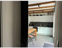 Moderne Küche mit weißen Oberschränken und schwarzer Arbeitsplatte, darunter ein integrierter Herd. Die Decke ist mit sichtbaren Holzbalken versehen, und ein hölzerner Esstisch mit Barhockern steht zentral im Raum.