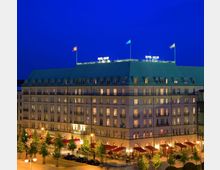 Ein mehrstöckiges Gebäude mit historischem Design und einer grünen Dachkonstruktion, beleuchtet in der Abenddämmerung. Es handelt sich um das "Hotel Adlon", mit großen Fenstern, Balkonen, rotem Vordach und einem belebten Eingangsbereich, umgeben von Bäumen und Straßenlaternen.