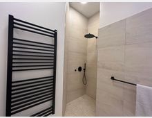 Minimalistisches Badezimmer mit großen cremefarbenen Fliesen, einer offenen Dusche mit Regenduschkopf und schwarzer Armatur. Links ein schwarzer Handtuchheizkörper, rechts eine Handtuchstange mit weißem Handtuch.