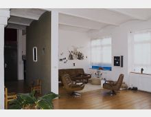 Wohnbereich in einem Loft mit Holzboden, weißen Wänden und großen Sprossenfenstern. Die Einrichtung umfasst braune Ledersessel, ein Sofa, einen flauschigen Teppich, Kunstwerke an den Wänden und Zimmerpflanzen.