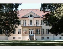 Gelbes, zweigeschossiges Herrenhaus mit roten Dachziegeln, symmetrischen weißen Sprossenfenstern und einer zentralen Eingangstreppe, umgeben von Rasen und Bäumen.