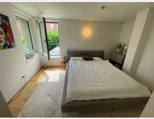 Helles, modern eingerichtetes Schlafzimmer mit Doppelbett, hellem Teppich und Holzfußboden. Links sorgen große, teils geöffnete Fenster für Tageslicht und Blick ins Grüne; rechts stehen eine Kommode und eine Wandlampe an einer schrägen Wandnische.