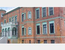 Backsteingebäude im historischen Stil mit grünen Fensterrahmen und dekorativen Elementen, darunter Rundbogenfenster und ein kleiner Vorbau mit verzierten Balustraden. Die Fassade ist symmetrisch gestaltet, und der Bau weist typische Merkmale der Backsteinarchitektur auf.
