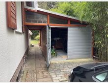 Seitlicher Hofbereich eines Wohnhauses mit schmalem gepflastertem Weg und einem angebauten Holzschuppen mit flachem Dach und teilweise geöffnetem grauen Schiebetor. Rechts stehen dichte Bambuspflanzen, im Vordergrund ist ein Teil eines geparkten Autos zu sehen.