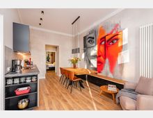 Offener Wohn- und Essbereich mit integrierter Küchenzeile, Holzfußboden und modernem Esstisch mit Stühlen unter schlanken Pendelleuchten. Auffällig ist ein großflächiges Wandgemälde mit einem Gesicht in Grau- und Rottönen, daneben ein Sofa und ein weißer Heizkörper.