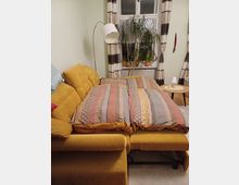 Wohnzimmer mit ausziehbarem gelbem Sofa, das als Bett hergerichtet ist, mit gemusterten Bettdecken in warmen Farben. Im Hintergrund ein großes Fenster mit gemusterten Vorhängen und einer Zimmerpflanze auf der Fensterbank, daneben eine Stehlampe und ein kleiner runder Tisch.