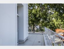 Eine helle Terrasse mit weiß gefliesten Böden und verzierten Metallgeländern, umgeben von grünen Bäumen. Im Hintergrund sind Dächer von Gebäuden sichtbar, und links befindet sich ein Abschnitt einer weißen Außenwand mit einer Glastür.