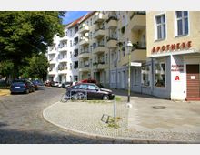 Straßenecke in einem Wohngebiet mit gepflastertem Gehweg, parkenden Autos und einer gelben Wohnhausfassade mit Balkonen. Auf der rechten Seite befindet sich eine Apotheke mit einer Beschilderung und Schaufenstern; daneben sind Straßenschilder sichtbar.