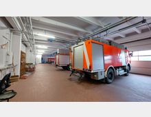 Innenansicht einer Feuerwehrgarage mit zwei geparkten roten Feuerwehrfahrzeugen. Der Raum hat eine helle Decke mit sichtbaren Lüftungsrohren, eine Ziegelbodenfläche und ein großes Garagentor mit Fenstern auf der rechten Seite.