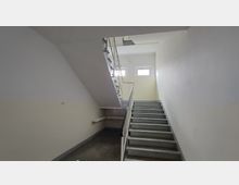 Treppenhaus in einem Gebäude mit weiß gestrichenen Wänden, Metallgeländer und zwei kleinen Fenstern, die natürliches Licht hereinlassen. Die Treppe führt nach oben und wirkt funktional und schlicht.