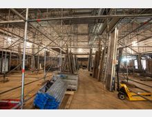Eine Baustelle in einem großen Innenraum mit zahlreichen Gerüsten aus Metallstrukturen, Holzplanken und Baugeräten, darunter ein gelber Hubwagen. Der Raum ist teilweise mit freiliegenden Ziegelwänden und provisorischer Beleuchtung ausgestattet.
