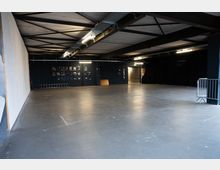 Große, leere Industriehalle mit glattem Betonboden, schwarzen Wänden und freiliegenden Stahlträgern unter der Decke. Mehrere Neonröhren und Kabeltrassen hängen an der Decke; rechts sind schwarze Vorhänge und ein Absperrgitter, hinten ein Durchgang zu einem weiteren Bereich.