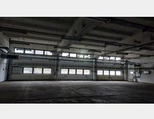 Leere Industriehalle mit hohen Decken, mehreren großen Fenstern an einer Wand und sichtbaren Rohrleitungen sowie Beleuchtungsanlagen an der Decke. Der Boden besteht aus Betonplatten, die Wände sind weiß gestrichen.