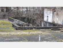 Verlassener, moosbewachsener Betonhof mit niedrigen, rechteckigen Mauern und einem runden Betonschachtdeckel. Rechts steht ein verwittertes Gebäude mit bröckelndem Putz und kleinem Fenster, im Hintergrund dichtes, überwuchertes Gehölz.