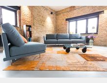 Wohnzimmer im industriellen Stil mit freiliegenden Ziegelwänden, grauem Sofa, modernem Sessel, Glastisch auf Rollen und einem großen orangenen Teppich. Im Hintergrund befinden sich große Fenster mit schwarzem Rahmen und links ein Regal mit Büchern.