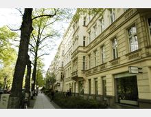 Straßenszene mit Altbau-Fassaden im historischen Stil, großen Fenstern und feinen Verzierungen; links eine Baumreihe entlang eines Gehwegs, rechts Schaufenster mit einem Schild „AGO Galerie“.
