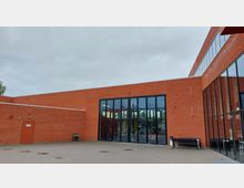 Außenansicht eines modernen Backsteingebäudes mit großen Glasfassaden und Fenstern, die den Innenraum sichtbar machen. Der Vorplatz ist gepflastert und ein Baum sowie Sitzbänke sind am Rand zu erkennen.