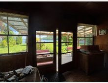 Dunkler Innenraum einer rustikalen Holzhütte mit großen Fenstern und einer Glastür zur überdachten Veranda. Draußen sind grüne Wiese, Sträucher und ein Blick auf ein Gewässer zu sehen, unter dem Vordach stehen rot-weiße Sitzbänke.