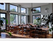 Helles Restaurant mit großen bodentiefen Fenstern, durch die viel Tageslicht einfällt. Der Raum ist mit Holzböden, schlichten Holztischen und dunkel gepolsterten Stühlen eingerichtet; Pflanzen ergänzen das gemütliche Ambiente.