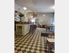 Ein gemütliches Café mit gemustertem Schachbrettboden, einer langen Theke aus hellem Holz mit Hockern und einer Blumenvase darauf. Im Hintergrund stehen Tische und Stühle, eine Fensterfront lässt Tageslicht herein, und ein Gast sitzt entspannt auf einem Sofa.