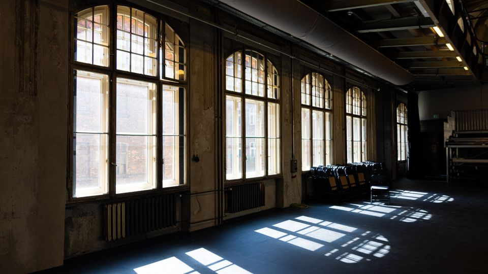Innenraum einer ehemaligen Industriehalle mit hohen Decken und großen, bogenförmigen Sprossenfenstern entlang einer Wand. Sonnenlicht fällt durch die Fenster und wirft rechteckige Schatten auf den Boden; an der Seite stehen Reihen von Stühlen und sichtbare Heizkörper unter den Fenstern.