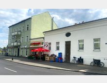 Straßenszene mit einem grün gestrichenen Wohnhaus mit Balkonen und einem angrenzenden weißen Gebäude mit dekorativem ovalem Fenster. Vor dem Gebäude befindet sich ein Spätkauf mit einem roten Sonnenschirm und Obst- und Gemüsekisten; daneben sind Fahrräder an der Wand abgestellt.