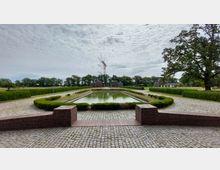 Großzügige Gartenanlage mit symmetrischem Wasserbecken, umrahmt von niedrigen Hecken und Pflasterwegen. Im Hintergrund stehen Backsteingebäude, Bäume und ein großer Funkmast vor einem leicht bewölkten Himmel.