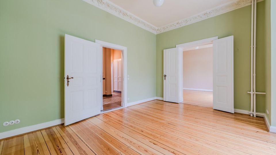 Leerer Altbau-Wohnraum mit grün gestrichenen Wänden, hellem Holzdielenboden und Stuckverzierungen an der Decke. Zwei weiße Doppeltüren führen in angrenzende Räume, rechts sind sichtbare Heizungsrohre an der Wand.