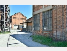 Industriegelände mit historischen Backsteingebäuden und großen Fensterflächen. Im Vordergrund verläuft ein Schienenstrang, umgeben von Gehwegen und Metallzäunen, unter klarem Himmel.