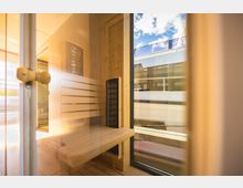 Holzverkleideter Saunaraum mit einer Sitzbank und einem schwarzen Heizkörper, durch eine Glastür mit Holzgriff sichtbar; im Hintergrund Blick nach draußen auf Balkongeländer und blauen Himmel.