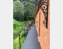 Ein Balkon aus Holzlatten an einem roten Ziegelgebäude mit blick auf eine grüne, bewachsene Umgebung und Bäume. Metallgeländer und dekorative Eisenelemente schmücken den Bereich, auf dem Holzstühle stehen.