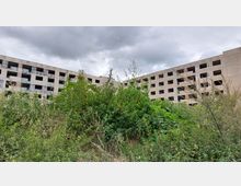 Ein verlassenes, mehrstöckiges Betongebäude mit offenen, unverglasten Fensteröffnungen, umgeben von dichtem, überwuchertem Grün und wildem Bewuchs. Der Himmel ist bewölkt und grau.