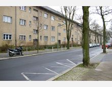 Straßenszene mit mehrstöckigem Wohngebäude aus beigefarbenem Putz entlang einer Baumreihe. Auf dem Gehweg sind ein Motorrad und geparkte Autos zu sehen, mit einer asphaltierten Fahrbahn im Vordergrund.