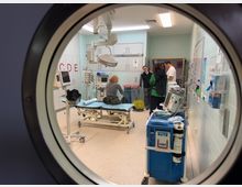 Blick durch ein rundes Fenster in einen sterilen Behandlungsraum mit blauen Fliesenwänden und medizinischen Geräten, darunter ein Behandlungsbett, Überwachungsmonitore und ein blauer medizinischer Wagen. Im Raum stehen mehrere Personen, die miteinander sprechen.