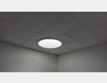 Decke eines Raumes mit grauen, rechteckigen Akustikpaneelen in einem Rautenmuster, in der Mitte eine große, runde Deckenleuchte mit weißem Licht.