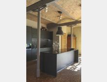 Moderne Küche mit schwarzen Einbau-Schränken und einer großen schwarzen Kücheninsel in der Mitte. Die Decke besteht aus Ziegelsteinen, gestützt durch industrielle Metallpfeiler, und der Boden ist aus Holzparkett.