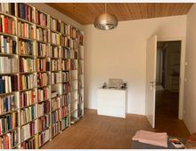 Ein Raum mit einer Holzbalkendecke, einer Bücherwand auf der linken Seite und einem hellen Holzboden. Rechts steht eine weiße Kommode vor einer weißen Wand, daneben eine geöffnete Tür, die in einen Flur führt.