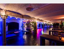 Großzügiger Raum mit industriellem Design, erkennbar an freiliegenden Backsteindecken und metallenen Säulen. Die Atmosphäre wird durch bunte, stimmungsvolle Beleuchtung, Holzakzente, dekorative Pflanzen und eine Bar im Hintergrund geprägt.