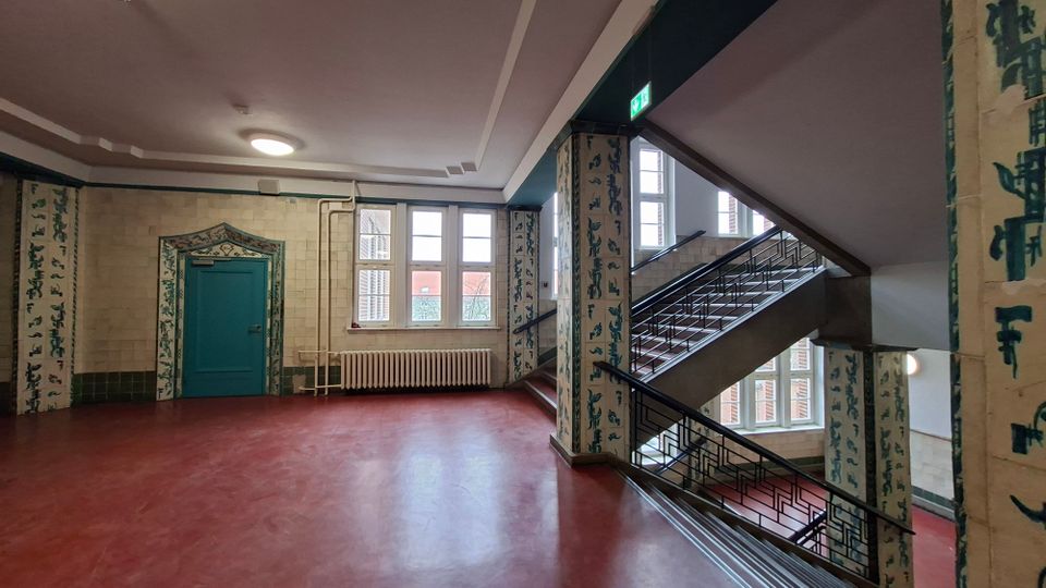 Innenansicht eines Treppenhauses in einem älteren Gebäude mit roten Linoleumböden, großem Treppenaufgang mit Metallgeländer und hohen Fenstern. Die Wände sind gefliest und mit vertikalen, blaugrünen Ziermustern sowie einer türkisfarbenen Tür mit dekorativem Rahmen gestaltet.