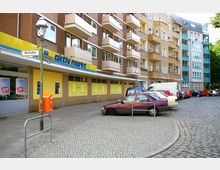 Straßenszene mit einem Wohn- und Geschäftshaus, dessen Erdgeschoss von einem gelben "aktiv markt"-Lebensmittelgeschäft eingenommen wird. Vor dem Gebäude befinden sich parkende Autos auf einem gepflasterten Platz, daneben weitere mehrstöckige Wohnhäuser.